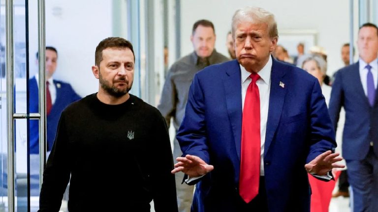 Donald Trump in a blue suit and red tie walking alongside Volodymyr Zelenskyy in a black sweatshirt through a modern hallway.