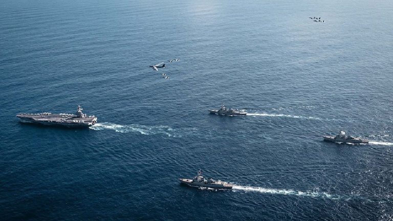 An aerial, high-angle view of a large naval fleet including an aircraft carrier, three escort ships, and a formation of military aircraft flying over a deep blue ocean.