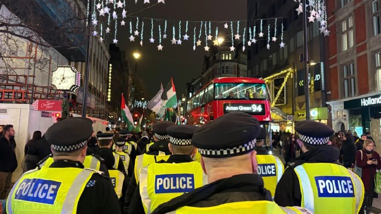 A line of UK police officers in yellow high-visibility vests facing a street with Palestinian flags and a red double-decker bus at night.