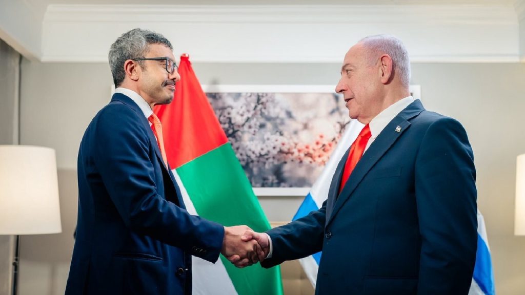 Two men in suits shaking hands indoors with national flags in the background.