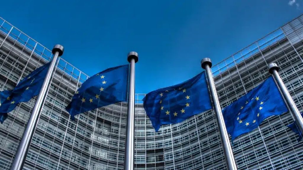 Several European Union flags waving in front of a modern curved glass building.