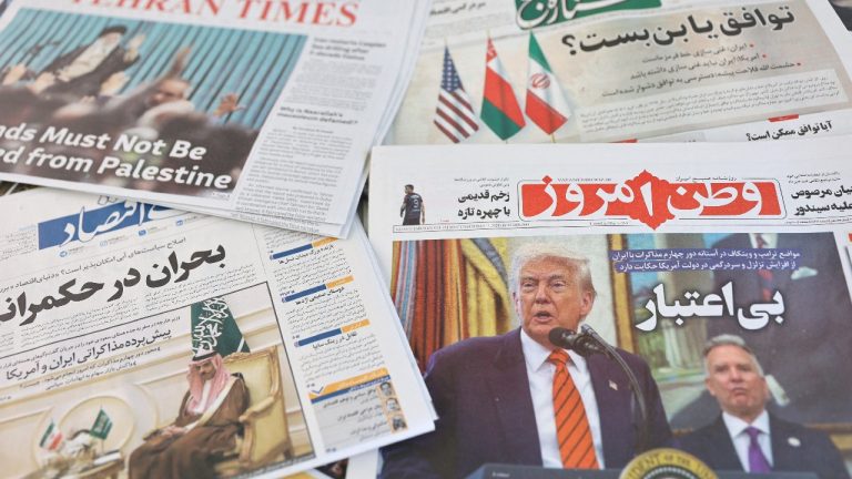 A collection of various Iranian newspapers spread out on a wooden table, featuring headlines in Persian and a prominent photograph of Donald Trump on one of the front pages.
