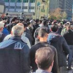 A large crowd of people walking down a city street, viewed from behind.