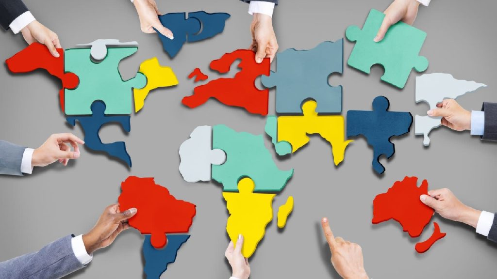 Hands of various people assembling a world map made of colorful puzzle pieces on a grey background.