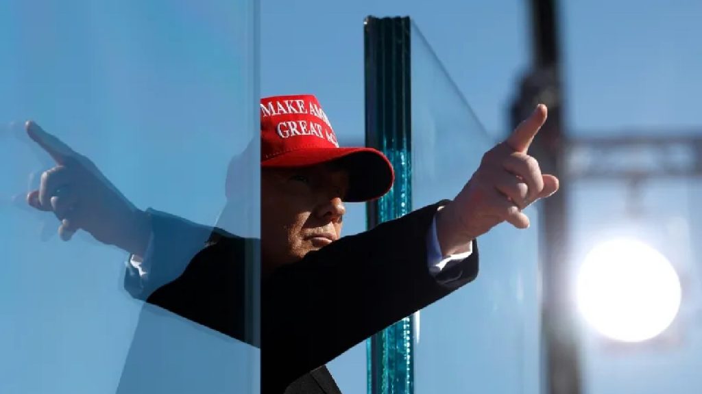 Donald Trump wearing a red "Make America Great Again" hat and pointing his finger through a glass pane.