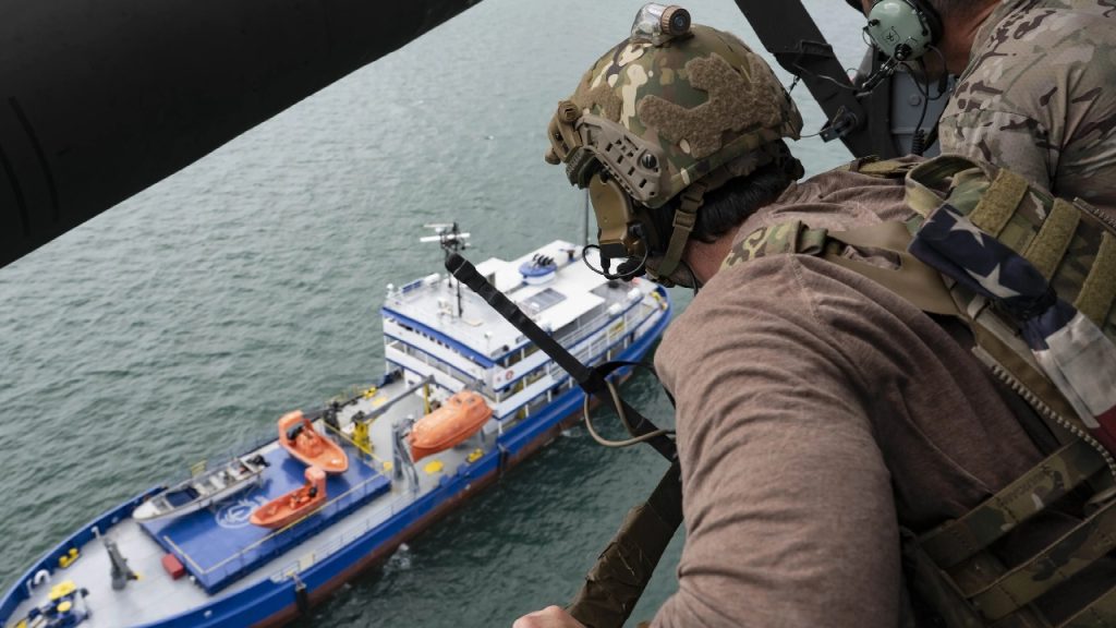 A soldier in military gear looking out from a helicopter at a blue and white ship on the water.