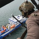 A soldier in military gear looking out from a helicopter at a blue and white ship on the water.