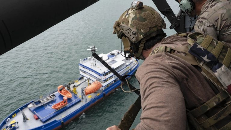 A soldier in military gear looking out from a helicopter at a blue and white ship on the water.