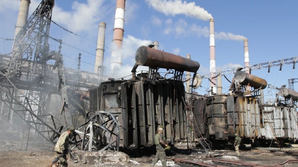 A wide shot of a heavily damaged industrial power plant in Ukraine, showing collapsed roofs, charred metal structures, and debris scattered across the site.