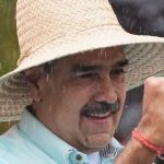 Close-up of a smiling man with a mustache wearing a wide-brimmed straw hat and a light blue shirt.