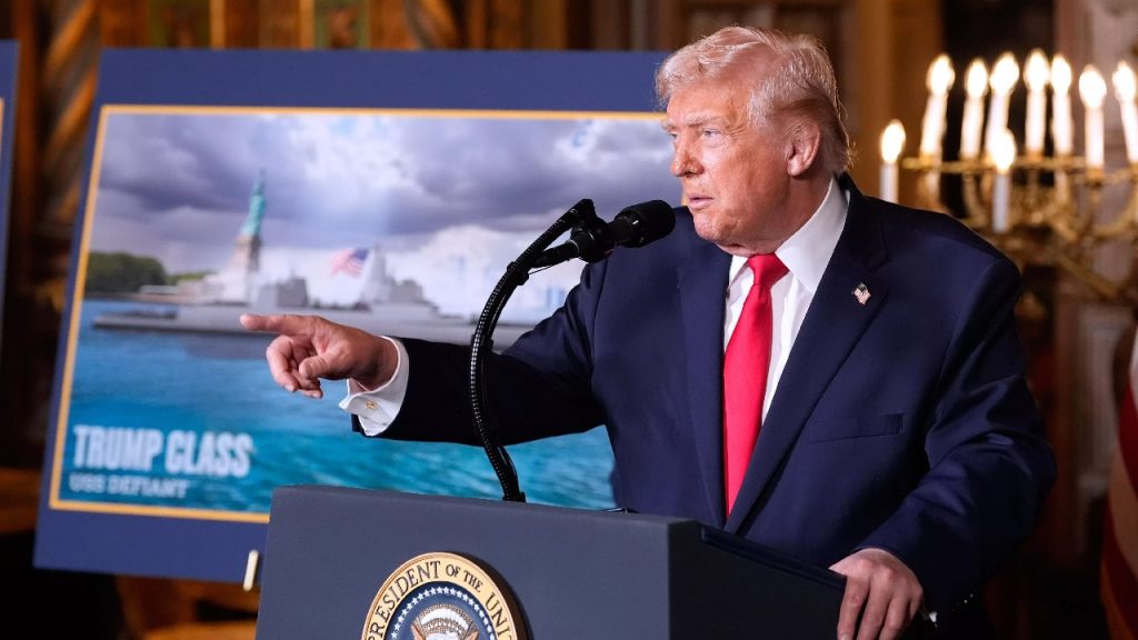 Donald Trump speaking at a podium with a large poster of a naval ship and the Statue of Liberty in the background.