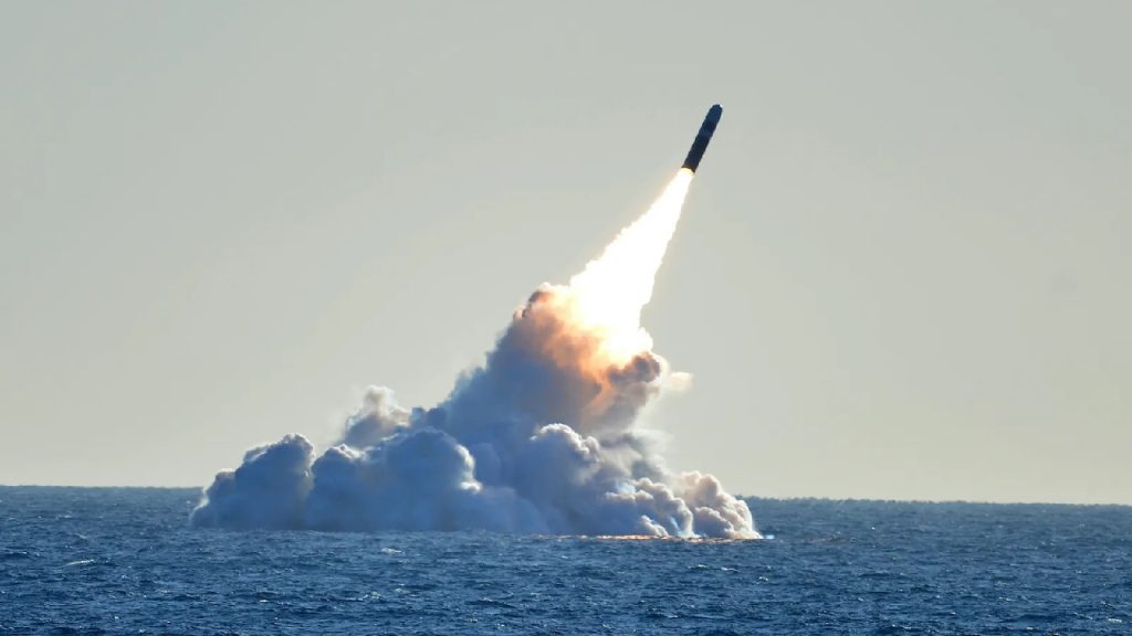 A dark missile launching vertically from the blue ocean with a large plume of white smoke and a bright fire trail at its base.