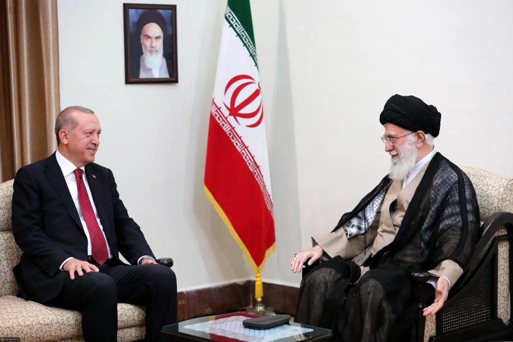 Two high-ranking political and religious leaders, Recep Tayyip Erdogan and Ali Khamenei, sitting in armchairs and smiling during a formal meeting, with the Iranian flag and a portrait of Ruhollah Khomeini in the background.