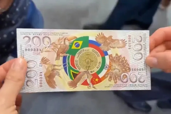 A pair of hands holding a sample banknote featuring the number "200" and a circular design in the center with the flags of Brazil, Russia, India, China, and South Africa, surrounded by illustrations of native animals like a toucan and a peacock.