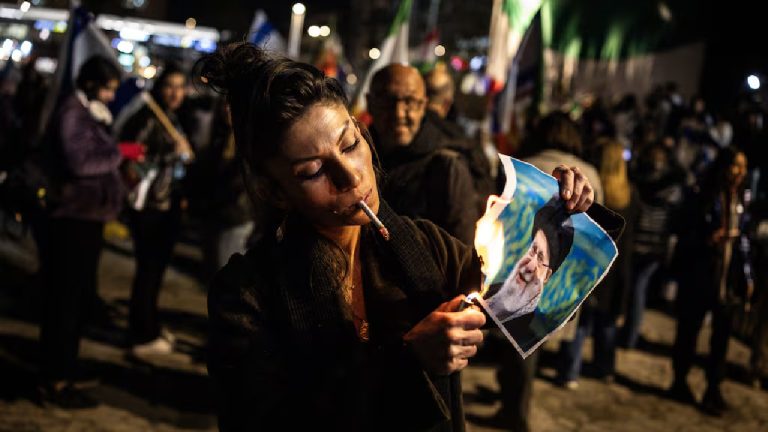 A woman in a dark coat sets fire to a printed portrait of a man with a white beard and black turban using a lighter.