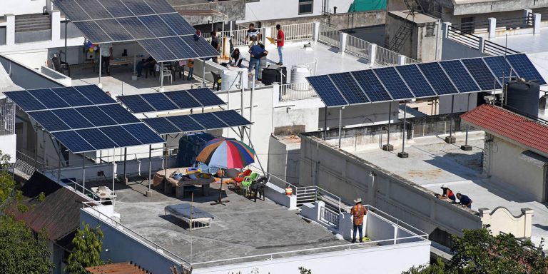A wide high-angle shot of a residential rooftop in a sunny urban area featuring multiple solar panel arrays and people gathered on the terrace.