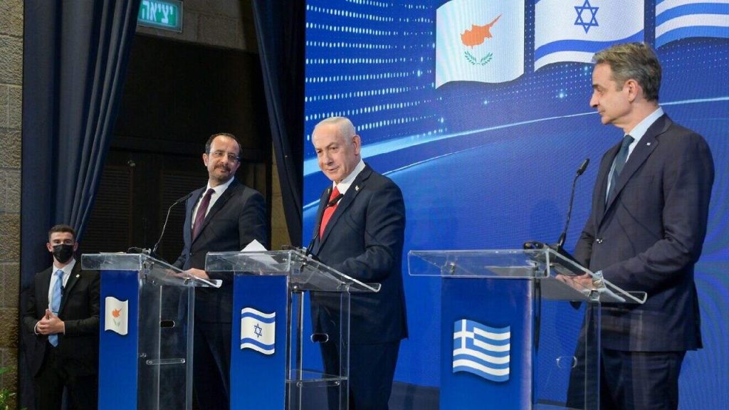 Three men in formal suits standing behind transparent lecterns featuring the flags of Cyprus, Israel, and Greece during a press conference.