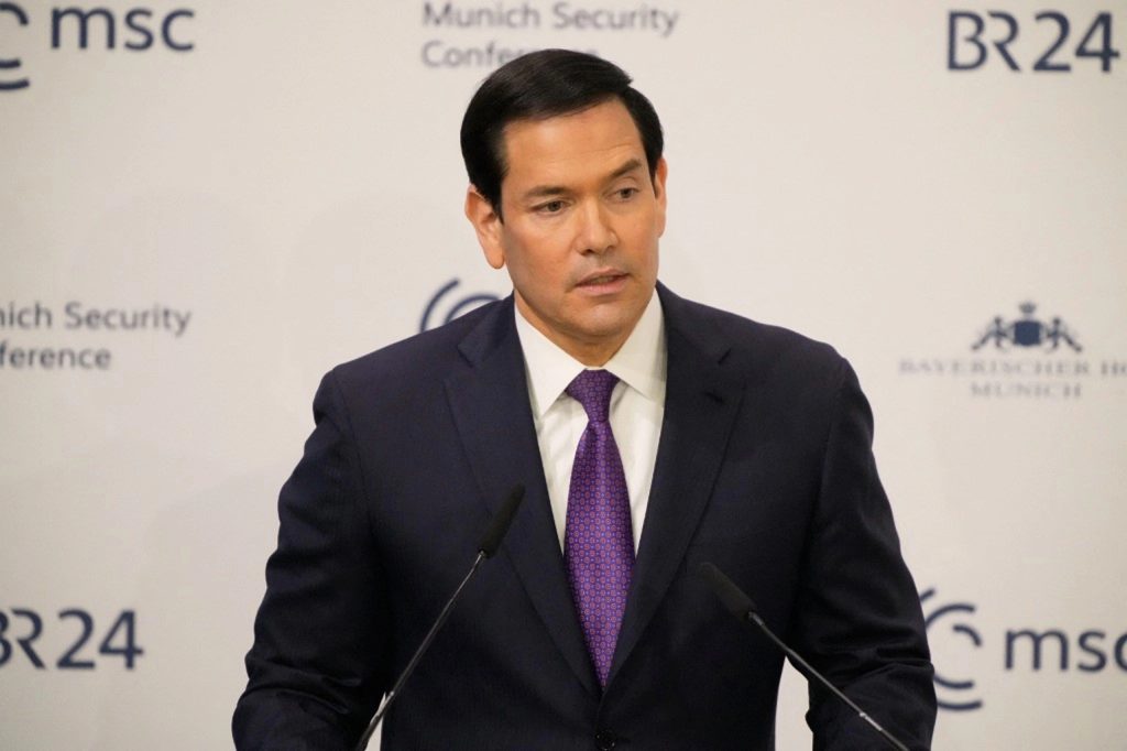 A medium shot of Marco Rubio speaking at a podium, wearing a dark navy suit and a patterned purple tie, with a white background featuring logos for the "Munich Security Conference" and "BR24."