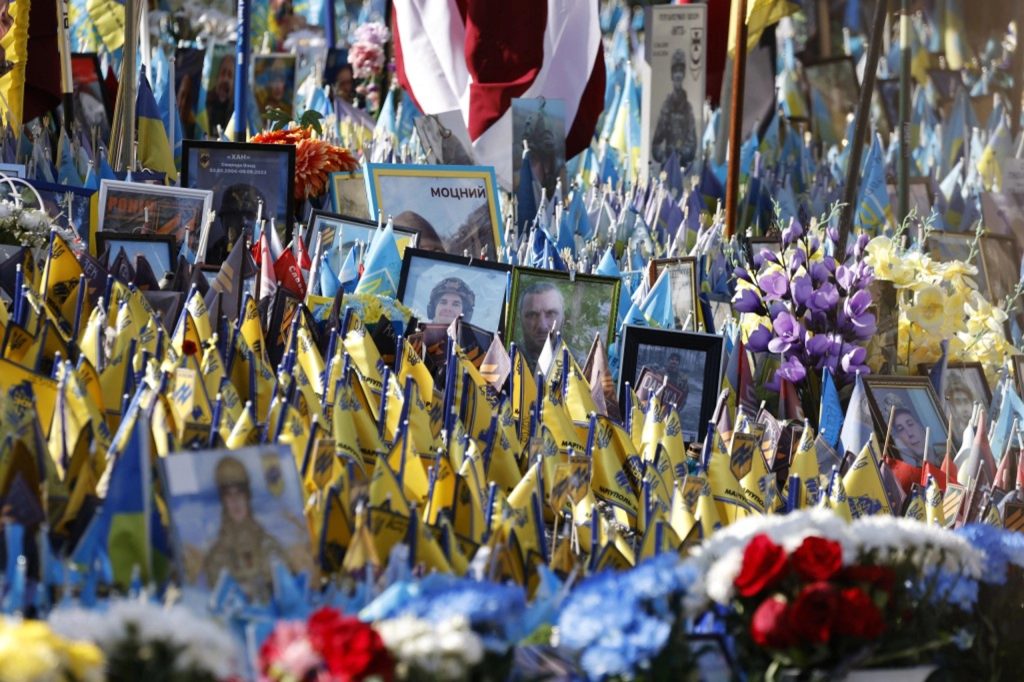 A dense memorial field filled with hundreds of small yellow and blue Ukrainian flags, interspersed with framed portraits of soldiers and colorful funeral flowers.
