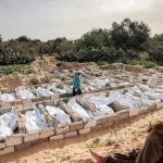 A young boy sitting on a dirt mound in the foreground, looking toward a large grid of open rectangular burial plots made of concrete blocks, where bodies wrapped in white shrouds are being placed by a man wearing a mask and a teal hoodie.