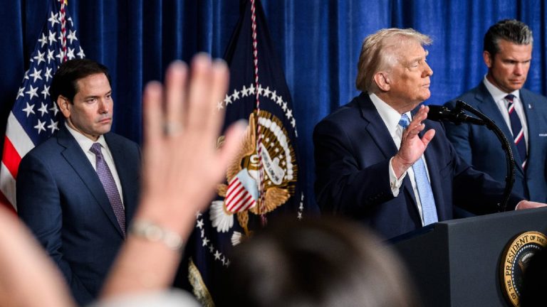 Donald Trump speaking at a podium with two men standing behind him and a hand raised in the foreground.