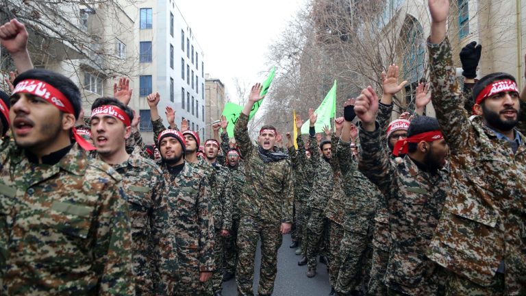 A large group of men in digital camouflage military uniforms marching on a city street, many wearing red headbands with white inscriptions and raising their hands or fists.