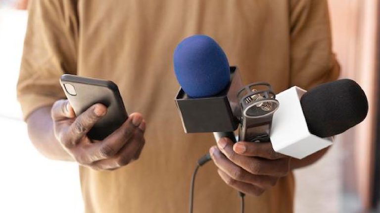A close-up of a person holding a smartphone in one hand and several news microphones with colorful foam covers in the other.