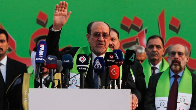 Nouri al-Maliki standing behind a podium with multiple news microphones, wearing a green sash and raising his right hand, surrounded by other men in suits against a green background with red Arabic text.