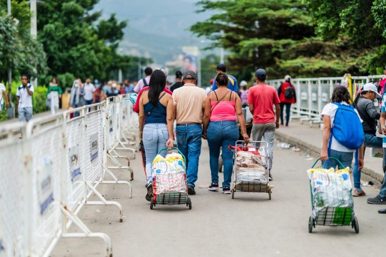 A medium shot from behind of people walking down a paved path, some pulling small metal carts filled with supplies, with white metal barriers on the left and lush green trees in the background.