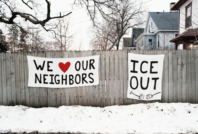Two white banners hanging on a wooden fence in a snowy residential neighborhood; the left banner reads "WE ❤️ OUR NEIGHBORS" and the right banner reads "ICE OUT" with an illustration of two brooms.