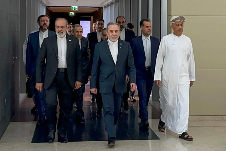 A group of men in formal suits, led by Abbas Araghchi and Majid Takht-Ravanchi, walking through a modern hallway, accompanied by an Omani official in traditional white attire and a patterned turban.