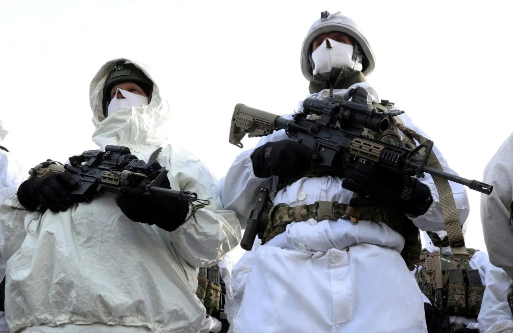 Two soldiers in full white winter camouflage gear, including white face masks and helmets, standing side-by-side and holding black assault rifles equipped with advanced scopes and tactical attachments.