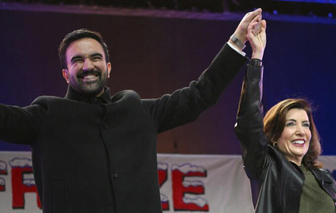 Zohran Mamdani and Kathy Hochul standing together on a stage, smiling and raising their joined hands in a gesture of unity and victory.
