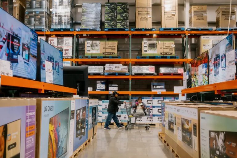 A man in a dark jacket pushing a shopping cart with a small child sitting inside it down an aisle in a large warehouse store, surrounded by high orange shelves stacked with large boxes of appliances and flat-screen televisions.