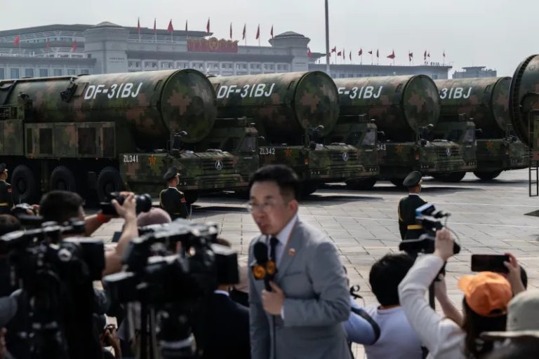 A line of large military trucks carrying camouflaged intercontinental ballistic missiles labeled "DF-31BJ" during a parade, with journalists and cameras in the foreground and a large building with red flags in the background.