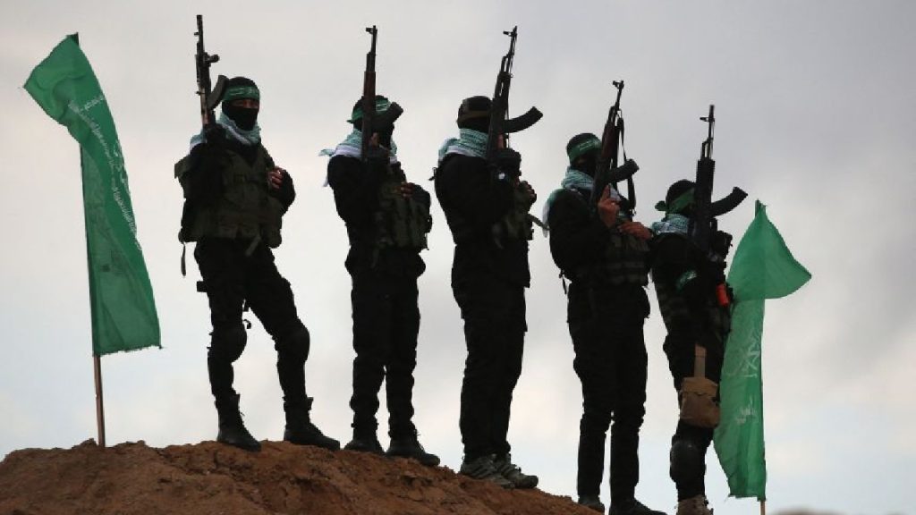 Five individuals in black tactical gear and face masks stand on a dirt mound holding rifles, flanked by two green flags.