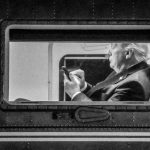 A black and white, grainy photograph showing Donald Trump from a side profile, viewed through the rectangular window of an aircraft, while he is looking down and using a smartphone.