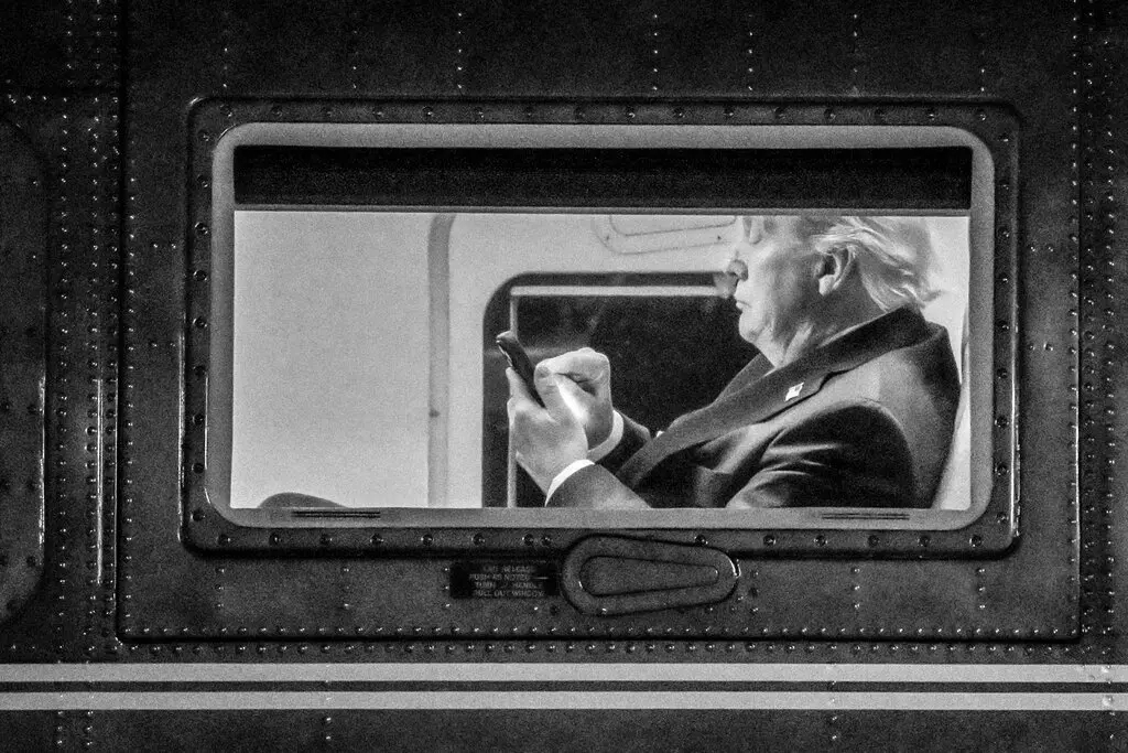A black and white, grainy photograph showing Donald Trump from a side profile, viewed through the rectangular window of an aircraft, while he is looking down and using a smartphone.