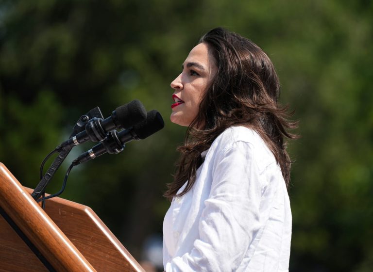 A side-profile close-up of Alexandria Ocasio-Cortez speaking at an outdoor podium with two microphones, wearing a white button-down shirt against a blurred green background of trees.
