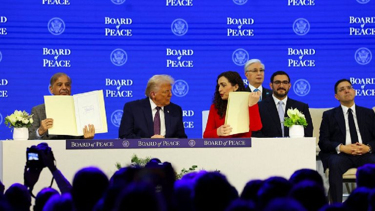 A group of men and one woman sit behind a long white table labeled "BOARD of PEACE" while two men hold up open folders showing documents.