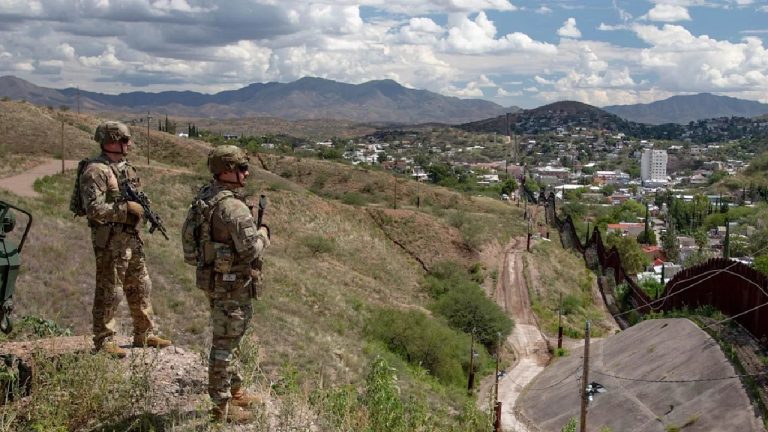 Two soldiers in camouflage uniforms and tactical gear standing on a rocky hill overlooking a border fence and a town in a valley.