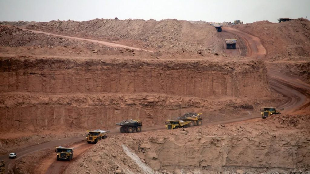 A wide-angle view of a massive open-pit mine with several large yellow haul trucks driving on dirt roads carved into the reddish-brown earth.
