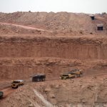 A wide-angle view of a massive open-pit mine with several large yellow haul trucks driving on dirt roads carved into the reddish-brown earth.