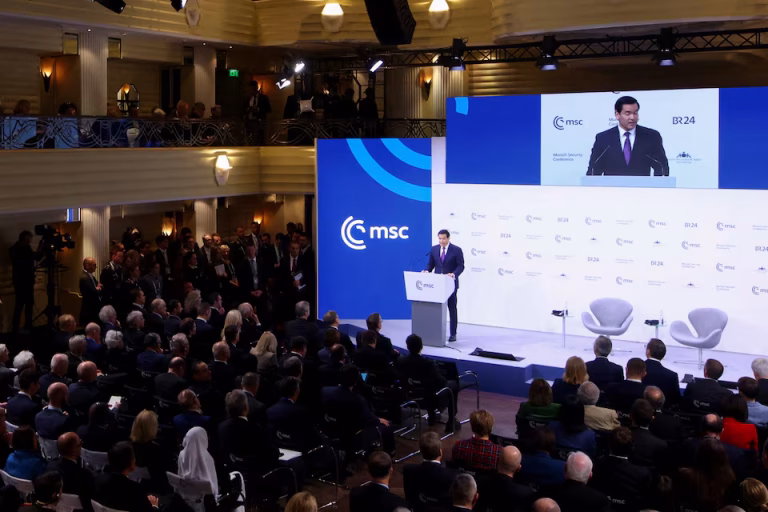 Marco Rubio speaking at a podium during the Munich Security Conference (MSC), with his image projected on a large screen above him and a seated audience in a grand hall.