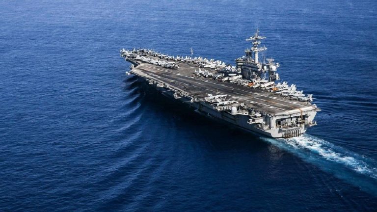 An aerial view of a large grey aircraft carrier sailing across a deep blue ocean, with several fighter jets parked on its flight deck.