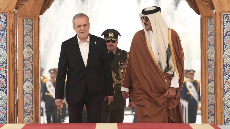 Two men in formal attire, one in a black suit and the other in traditional Middle Eastern clothing, walking together on a red carpet.