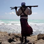 A person seen from behind, carrying a rifle across their shoulders and wearing ammunition belts, looking at the sea.