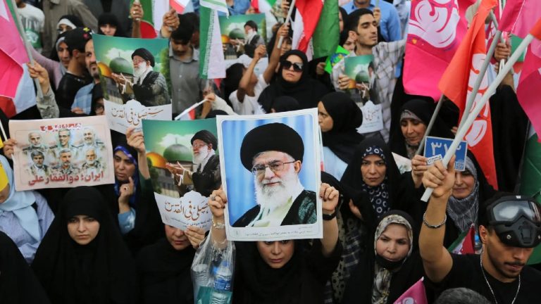 A large crowd of people holding up posters with a portrait of a bearded man in a black turban and glasses.