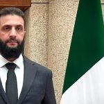 A medium shot of a man with a dark beard, wearing a grey suit and black tie, standing in front of a flag and a patterned wall.