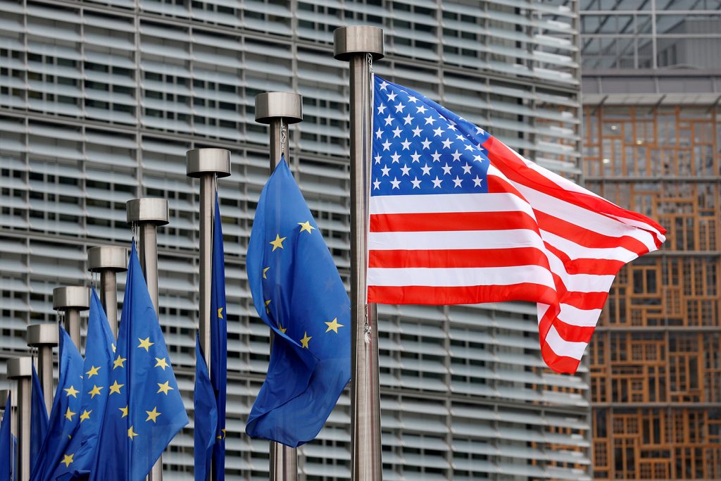 A close-up view of several silver flagpoles in a row, with multiple European Union flags and one prominent United States flag waving in front of a modern glass office building.